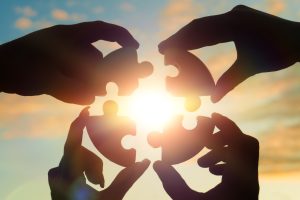 Four hands holding puzzle pieces against a bright sky, symbolizing teamwork and collaboration. Adobe Stock Image