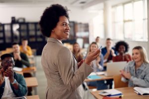 A woman stands in front of a classroom full of students sitting at desks. Adobe Stock
