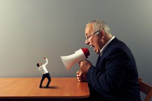 An artistic depiction of a very large man sitting at a table yelling into a megaphone at a very small person standing on the table with his hands raised. Adobe Stock