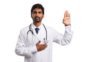 Person in a white lab coat with a stethoscope, raising one hand and placing the other on their chest, symbolizing a pledge or ethical commitment in the medical field. Adobe Stock