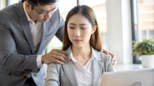 Two professionals in an office; one standing with his hands on the seated woman’s shoulders. The seated woman’s expression looks unassured of the physical touch. Adobe Stock