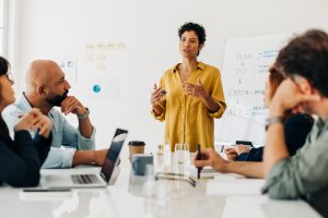 Person presenting to four seated colleagues in a meeting room. A white board in the background says Plan, do check, act. Adobe Stock