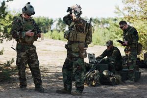 Four individuals in military camouflage gear conducting field operations with binoculars, maps and communication equipment in a wooded areas. Adobe Stock