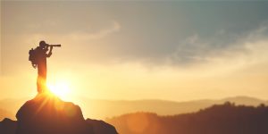 Silhouette of a person on a rocky peak using a monocular with mountains in the background during a sunrise or sunset. Adobe Stock